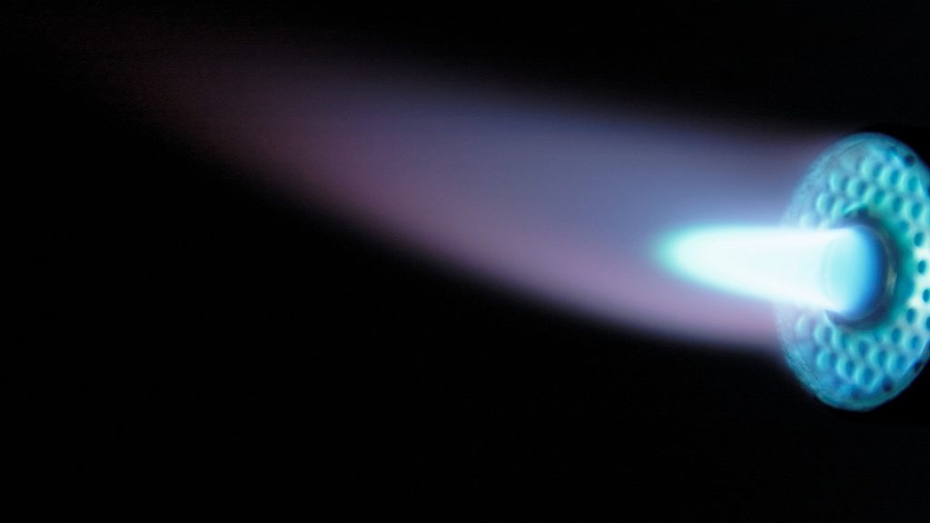 黒背景に映える青と紫の高速ジェット火炎（フレーム）。溶射材料を溶融・加速させ、マッハの速度で基材へ衝突させて被膜を形成するプロセスを表現。