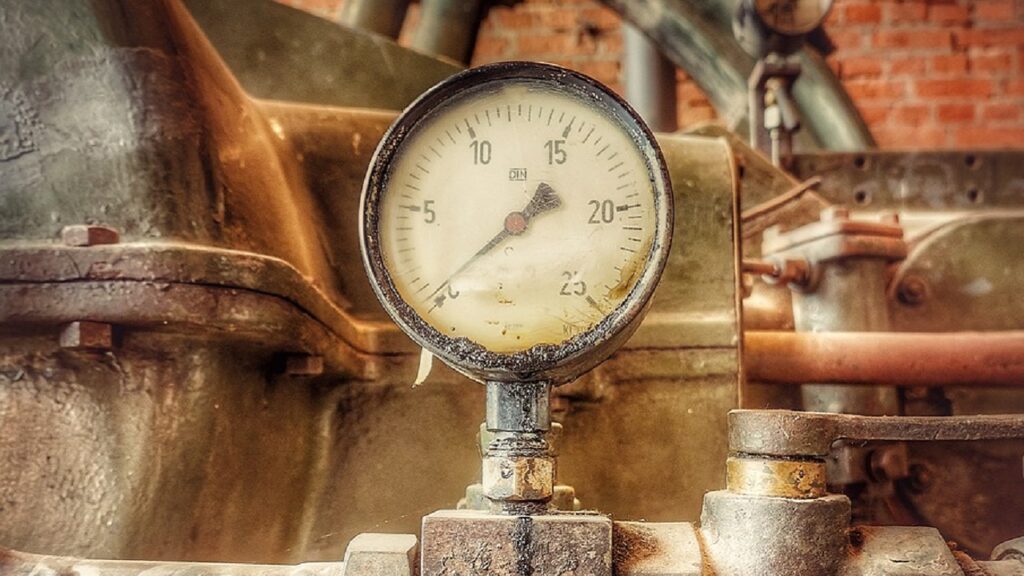 工場の配管に取り付けられた、経年変化で見づらくなった古い圧力計の写真。故障や視認性低下による交換の必要性と、グリセリン封入やネジ形状選定の重要性を象徴。