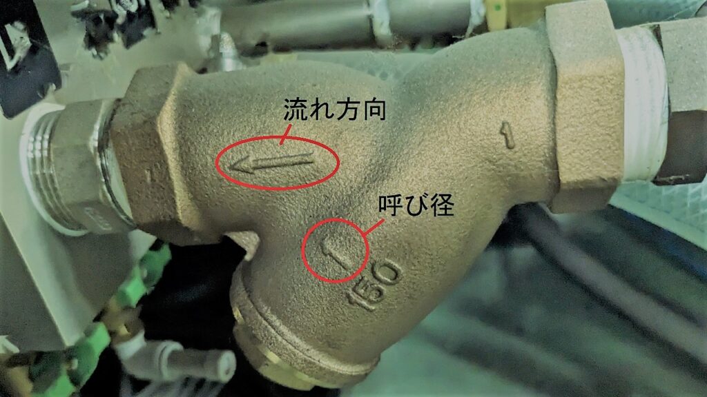 Y型ストレーナの表面に刻印された呼び径1（25A）と、流体の流れる方向を示す矢印の解説写真。